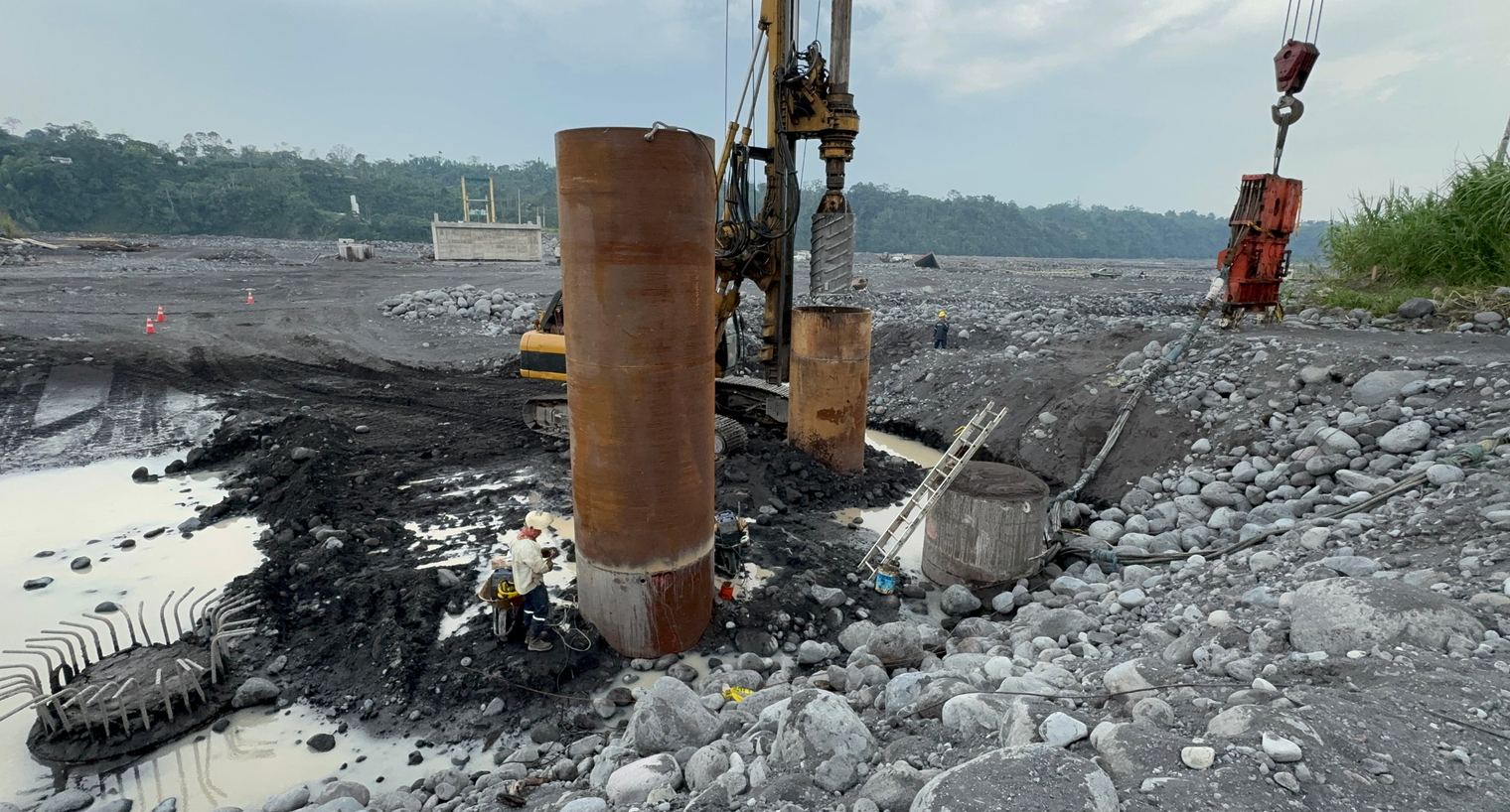 Steel pile casings on river gravel, with a rotary drilling rig behind and a vibratory hammer hanging from a crane